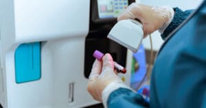 Gloved hands place a small blood sample vial into a clinical analyzer for testing.