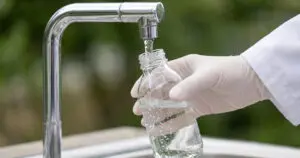 Gloved hand filling a small glass bottle with water from a laboratory faucet.