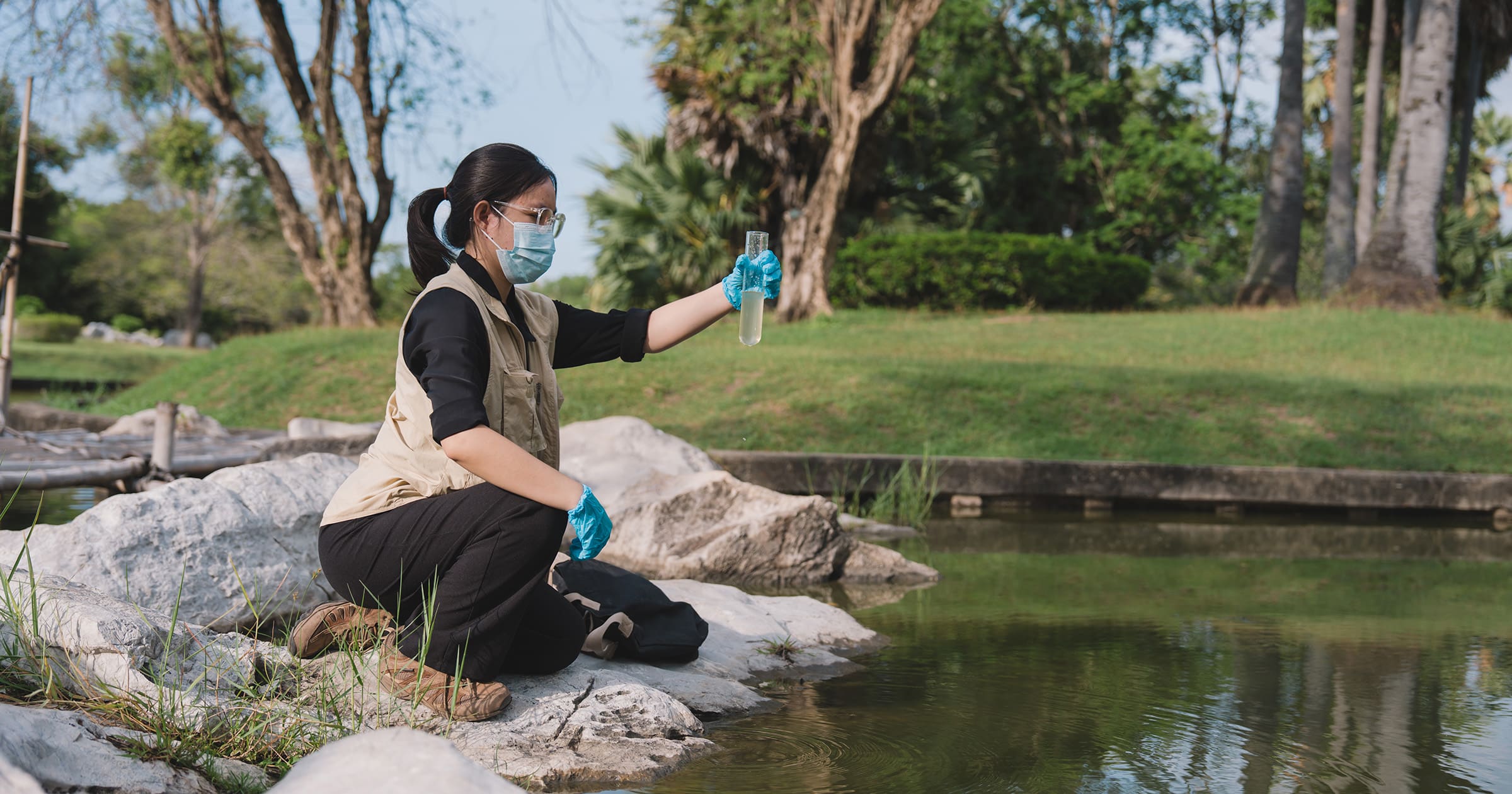 Person in mask and gloves kneels on rocks by a pond, holding a test tube for water sampling in a park.