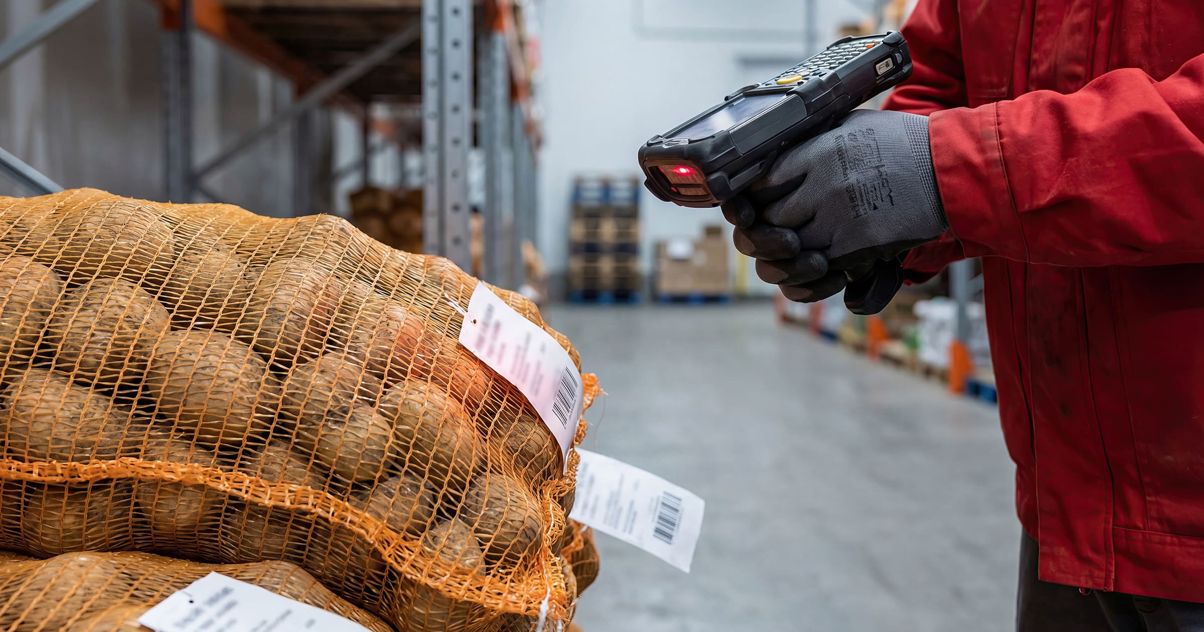 Warehouse worker in red protective clothing scans a barcode on a mesh bag with a handheld scanner by stacked sacks.