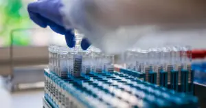 Gloved hand placing a test tube into a rack of labeled vials in a laboratory.