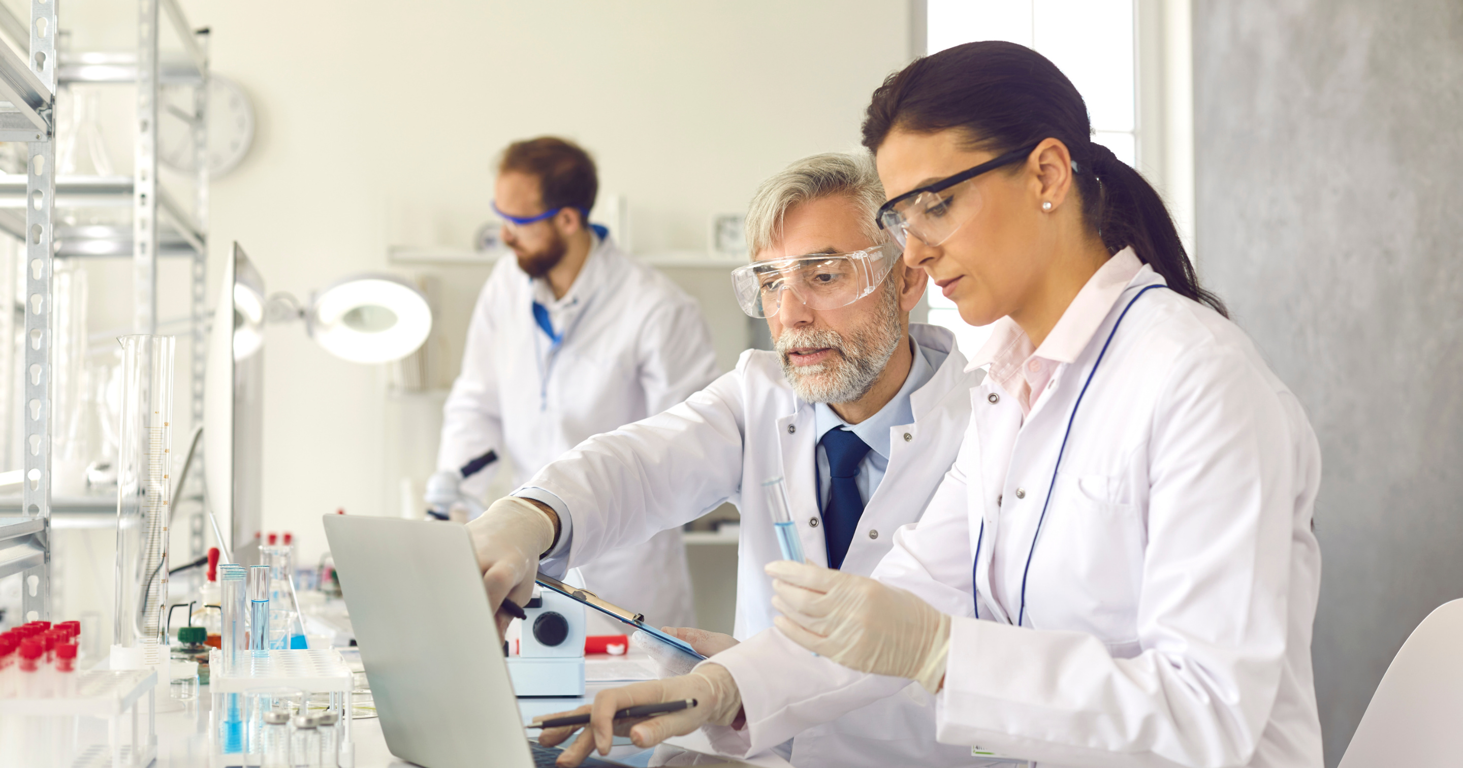 A senior scientist and a younger colleague reviewing results together on a laptop at a lab bench.
